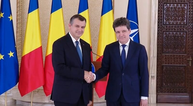 Two men in dark suits shake hands in a formal room, with EU, Romania, Ukraine, and NATO flags behind them.