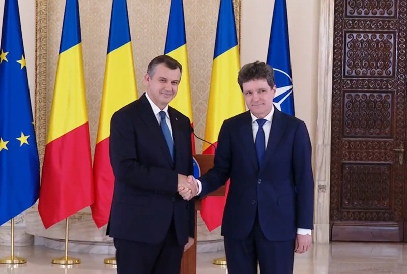 Two men in dark suits shake hands in a formal room, with EU, Romania, Ukraine, and NATO flags behind them.