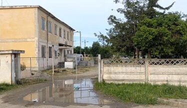 Two-story yellow building behind a chain-link gate on a muddy driveway with a large puddle and yard with trees to the right.