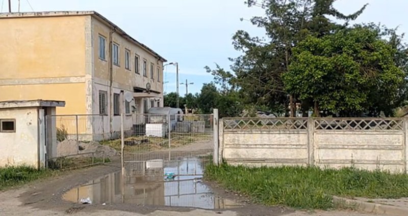 Two-story yellow building behind a chain-link gate on a muddy driveway with a large puddle and yard with trees to the right.