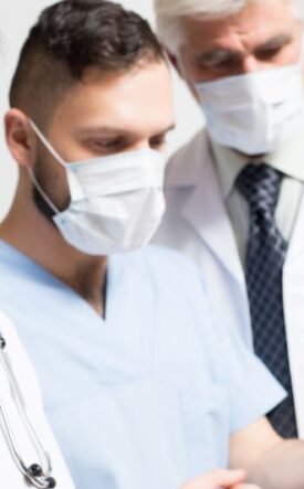 Three healthcare professionals wearing masks and white coats look at a tablet together, discussing patient care.