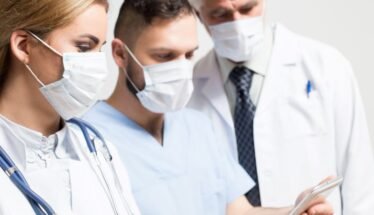 Three healthcare professionals wearing masks and white coats look at a tablet together, discussing patient care.