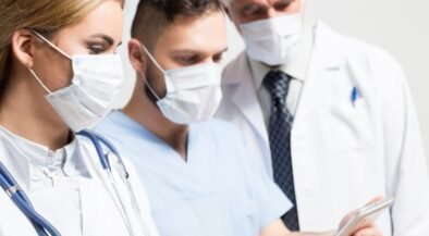 Three healthcare professionals wearing masks and white coats look at a tablet together, discussing patient care.