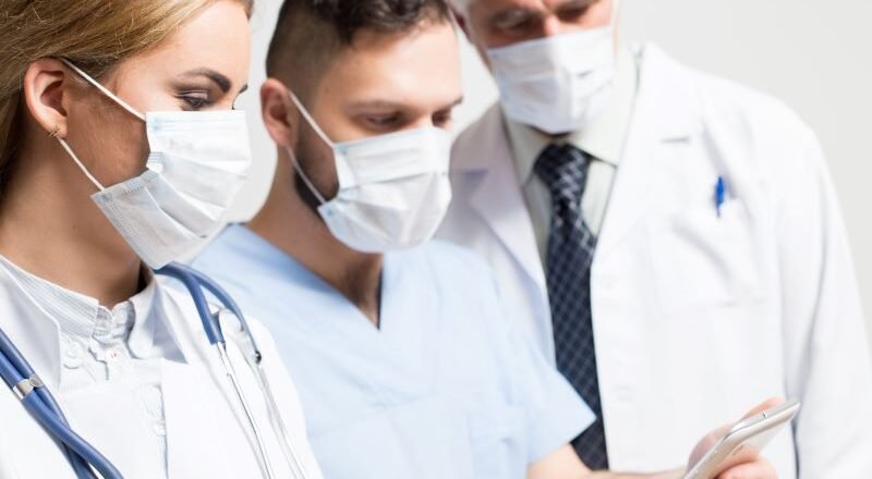 Three healthcare professionals wearing masks and white coats look at a tablet together, discussing patient care.