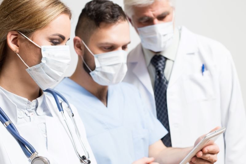 Three healthcare professionals wearing masks and white coats look at a tablet together, discussing patient care.