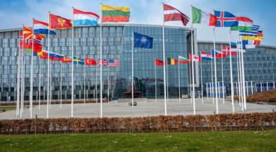 Modern glass-front building with row of flagpoles displaying numerous international flags in front of the plaza under a blue sky