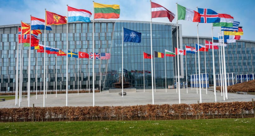 Modern glass-front building with row of flagpoles displaying numerous international flags in front of the plaza under a blue sky