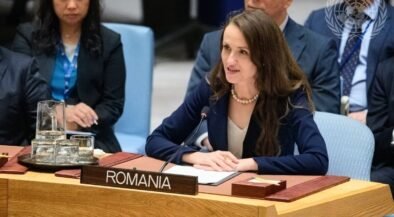 Romanian delegate speaking at a UN meeting, seated behind a Romania nameplate with a microphone in front of her.