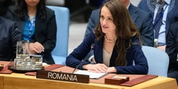 Romanian delegate speaking at a UN meeting, seated behind a Romania nameplate with a microphone in front of her.