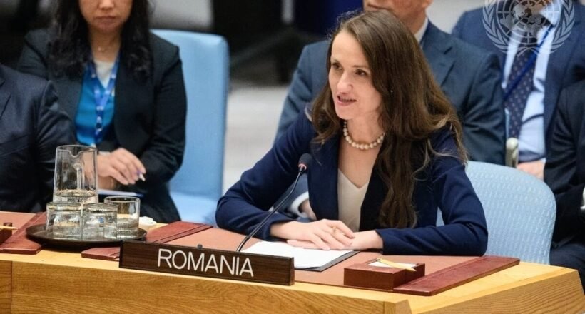 Romanian delegate speaking at a UN meeting, seated behind a Romania nameplate with a microphone in front of her.