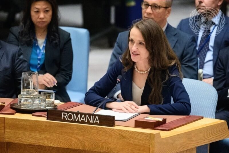 Romanian delegate speaking at a UN meeting, seated behind a Romania nameplate with a microphone in front of her.