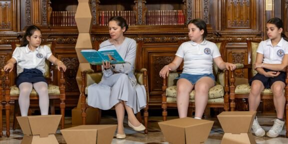 Teacher reads a blue book to four girls in white uniforms seated in an ornate wood library; cardboard art sculptures on the floor in front.