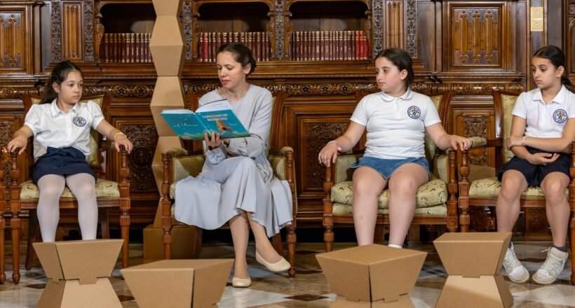Teacher reads a blue book to four girls in white uniforms seated in an ornate wood library; cardboard art sculptures on the floor in front.