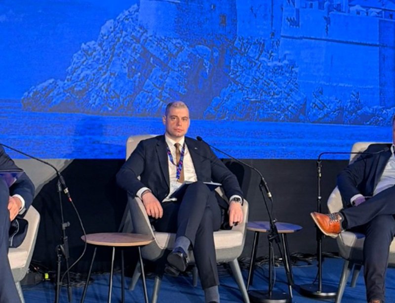 Man in a dark suit seated on a panel stage with a blue backdrop showing rocks and a coastline, microphones nearby.