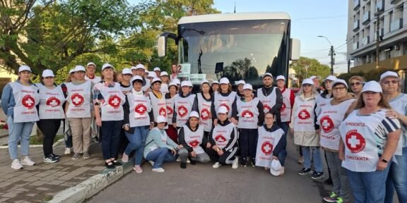 Group of Red Cross volunteers wearing white vests with red cross logos posing in front of a bus on a sunny street.
