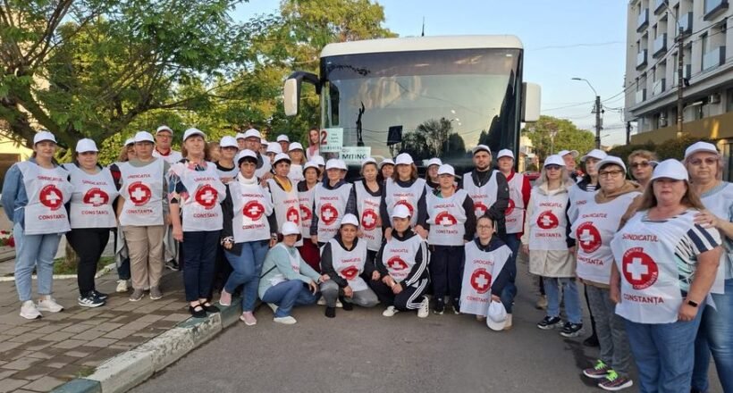 Group of Red Cross volunteers wearing white vests with red cross logos posing in front of a bus on a sunny street.