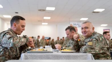 Three soldiers in camouflage sit at a table, eating in a bright mess hall with other service members in the background.