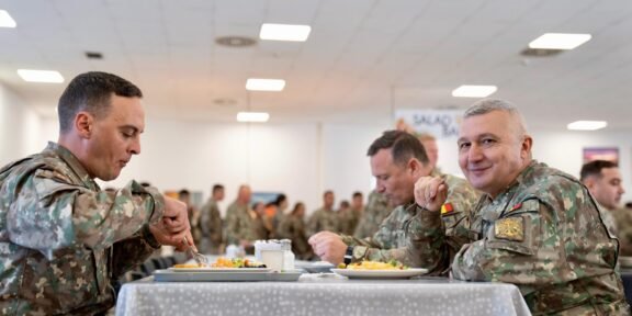 Three soldiers in camouflage sit at a table, eating in a bright mess hall with other service members in the background.