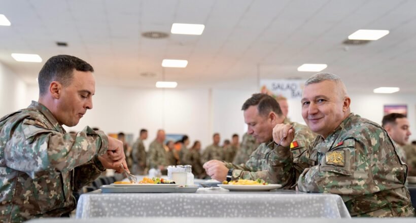 Three soldiers in camouflage sit at a table, eating in a bright mess hall with other service members in the background.