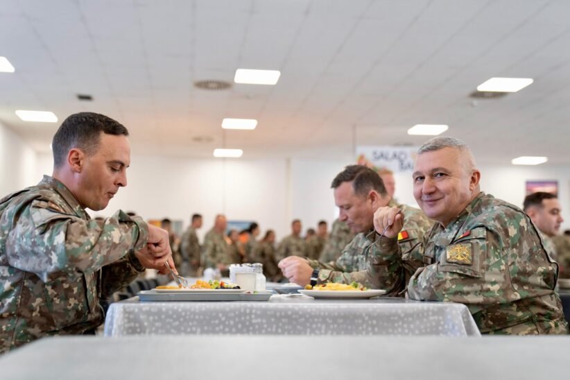 Three soldiers in camouflage sit at a table, eating in a bright mess hall with other service members in the background.